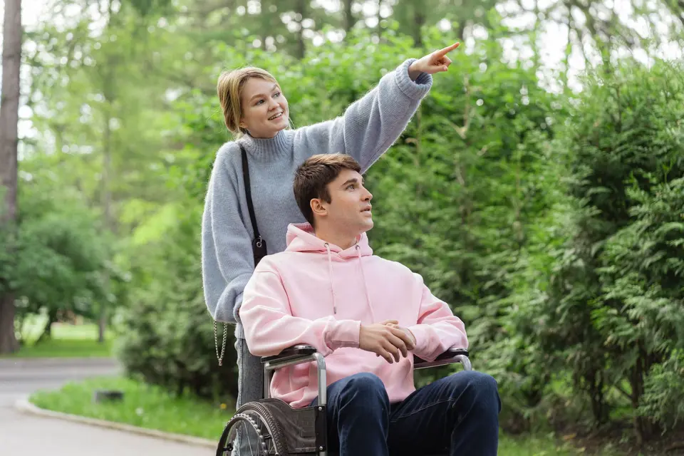 Een begeleider duwt een jongeman in een rolstoel door het park en wijst iets aan