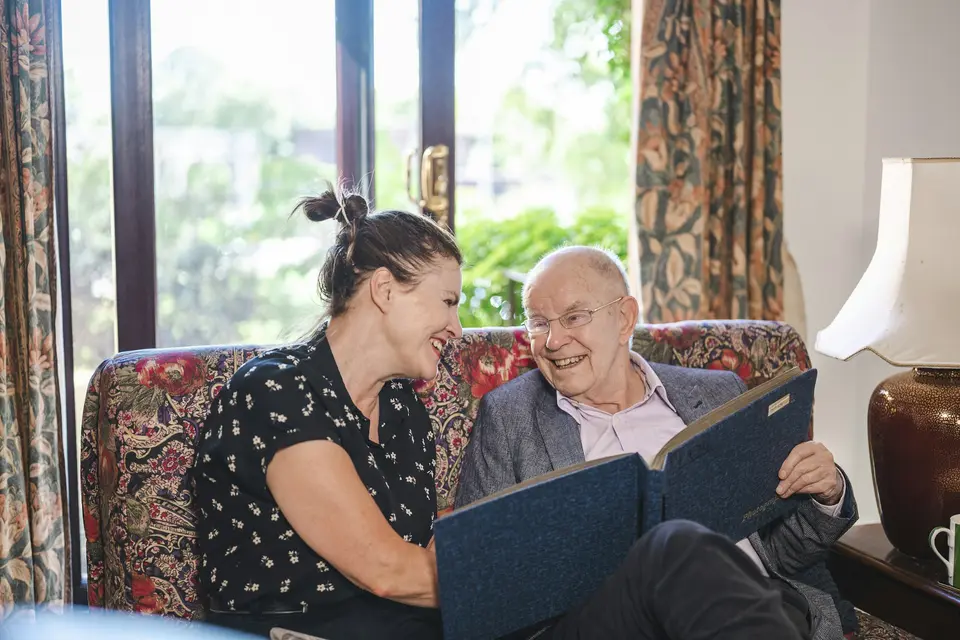 Een vrouw en een oudere man bekijken samen een fotoalbum op de bank in de woonkamer