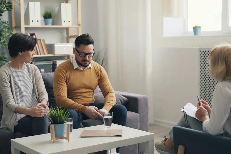 Een vrouw met een notitieblok voert een kennismakingsgesprek met een man en een vrouw in hun woonkamer