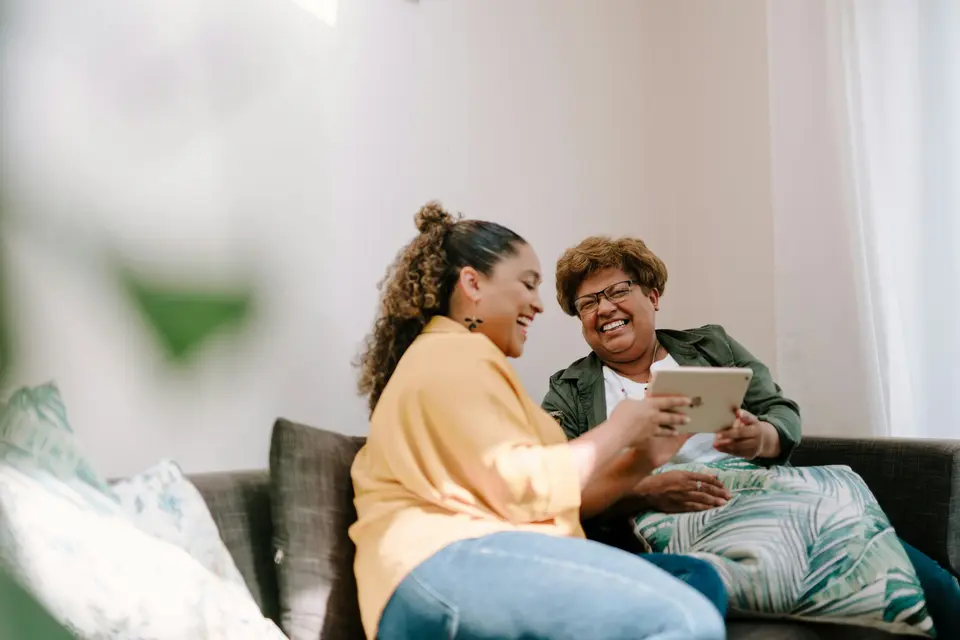 Een jonge vrouw laat een lachende oudere vrouw iets zien op een tablet terwijl ze samen op de bank zitten